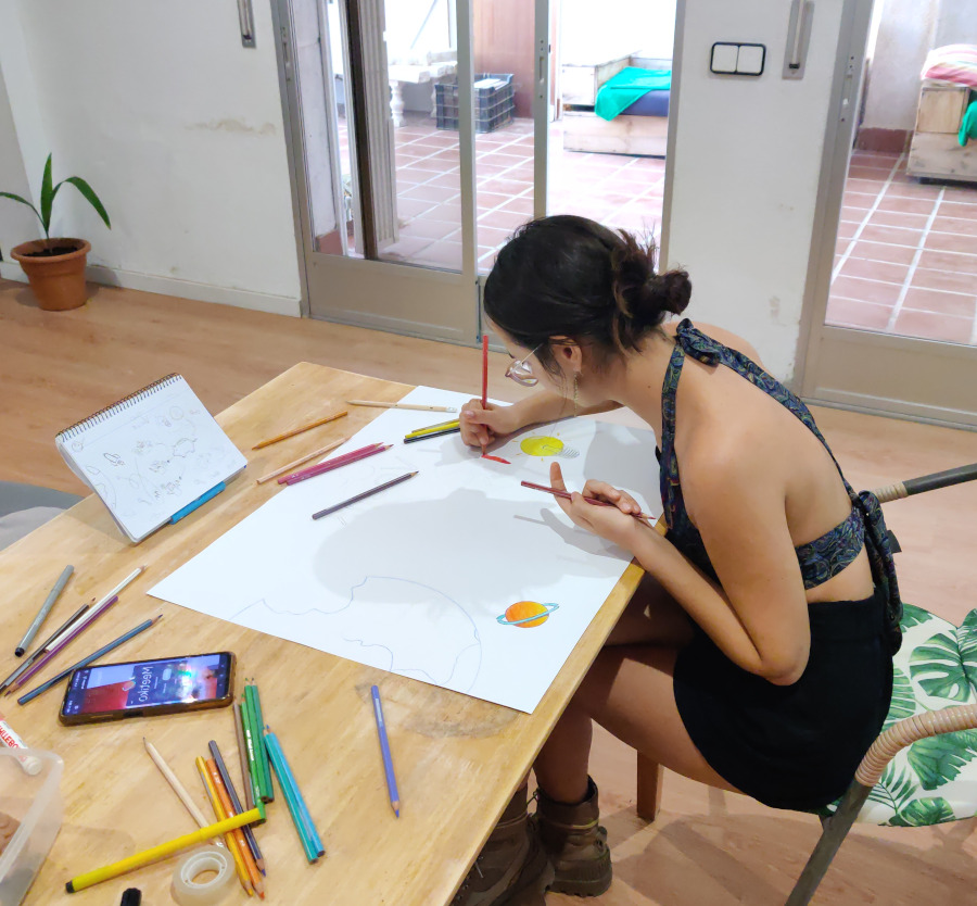 Photo of Elena in a room painting a poster board on a wooden table. You can see scattered crayons and the Meetiko logo painted on it.